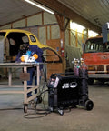 Person welding with Elite 200 Amp All In One Multi Process AC DC Welder MP200i in workshop, MIG Stick Ready, next to vintage car.