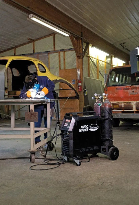 Person welding with Elite 200 Amp All In One Multi Process AC DC Welder MP200i in workshop, MIG Stick Ready, next to vintage car.