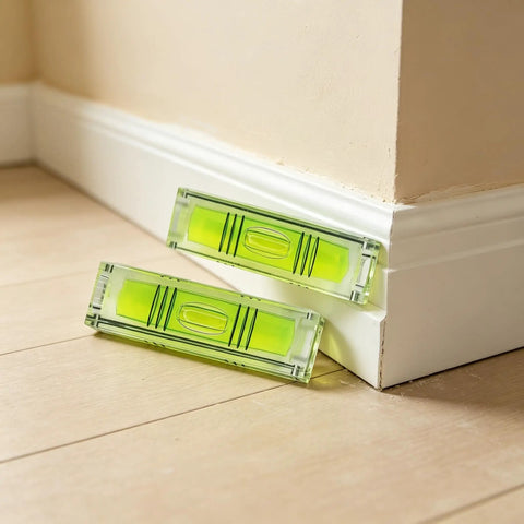 2 Pack Magnetic Mini Bubble Level Tool 2 Inch, Small Pocket resting against a wall corner on wood flooring.