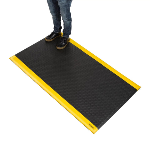 Industrial 15/16 Inch Extra Thick Anti Fatigue Floor Mat Built For Heavy Duty Jobs, Diamond Plate Non Slip Ergonomic Standing Mat In Black With Yellow Edge, Perfect For Workbench, Shop, Kitchen, Warehouse 3 By 5 Feet with person standing on it.