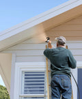 A painter using the Pro Grade High Pressure Electric Airless Paint Sprayer on a house's exterior siding while on a ladder.