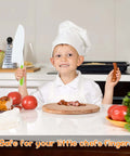 Front view of a child chef wearing a white hat, holding a green plastic toddler-safe kitchen knife over a cutting board.
