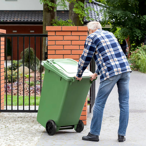 Person moving green garbage can with black wheels, demonstrating Garbage Can Wheels Replacement With Metal Hollow Shaft And Small Rubber Wheels 19.68 X 7.28 X 3.93 Inches Quiet Trash Can Pulley For Easy Installation And Efficient Waste Collection in a garden setting.