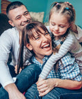 Happy family on a living room couch; mother laughs as daughter hugs from behind.