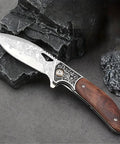 Dope Damascus Steel Folding Pocket Knife With 3.46 Inch Drop Point Blade, rosewood handle, on black rocks.