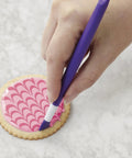 Hand using Top Shelf Cookie Decorating Tools Set Featuring Etching Wand on pink decorated cookie.