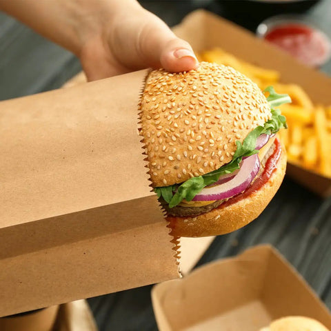 Hand holding burger in 100 Pack Brown Kraft Paper Lunch Bags With 4lb Capacity, 5 By 2.95 By 9.45 Inches Fold Top Disposable Bags