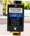 Pedestrian crossing signal with wait button, covered by round button protector, on a street post, outdoors.