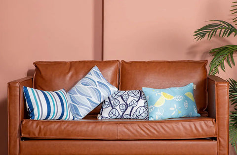 Four decorative pillows with various blue patterns on a brown leather sofa against a peach wall.