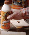 Hand holding Wood Glue For Indoor And Outdoor Projects, 18 Ounce Bottle, Natural Color, 2 Pack, Quick Clamp Time, on wooden table.