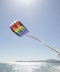 Rainbow Stripes 5.0 Parafoil Kite for all ages flying high above a beach, showcasing its bright colors and long tail against a clear sky.