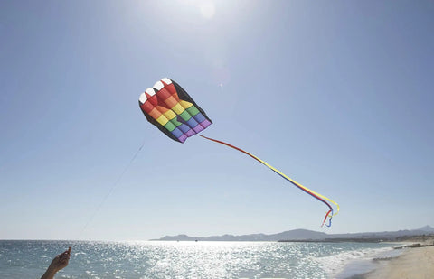 Rainbow Stripes 5.0 Parafoil Kite for all ages flying high above a beach, showcasing its bright colors and long tail against a clear sky.