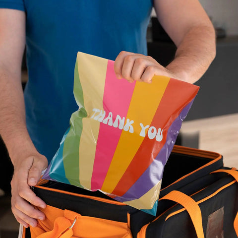 Person holding Rainbow Color Waterproof Poly Mailer Bags 100 Count Self Sealing Shipping Envelopes For Clothes above an orange bag.