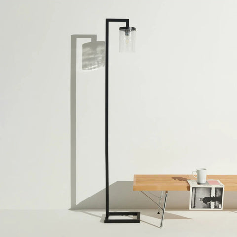 Modern black floor lamp next to a wooden table with books and a white cup, under natural light casting shadows on a white wall.
