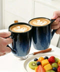 Two hands holding 14oz Ceramic Coffee Mugs Set Of 6 With Easy Grip Handle In Ice Snow Blue Glaze With Star Blue Accents And Gold Rim over a table.
