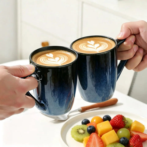 Two hands holding 14oz Ceramic Coffee Mugs Set Of 6 With Easy Grip Handle In Ice Snow Blue Glaze With Star Blue Accents And Gold Rim over a table.