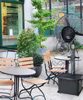 Rock Solid Outdoor Misting Oscillating Fan Featuring Indoor Cooling Pedestal Design in a patio setting with tables and chairs.