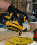 Person using a random orbital sander with 135 Piece 5 Inch Sanding Discs Set Eight Hole Round Hook And Loop Gold Pro Grade Grit on wood.