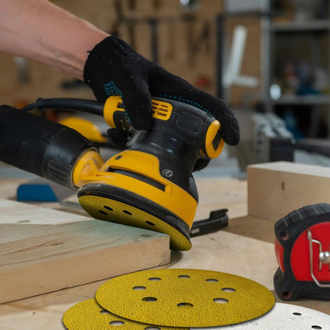 Person using a random orbital sander with 135 Piece 5 Inch Sanding Discs Set Eight Hole Round Hook And Loop Gold Pro Grade Grit on wood.