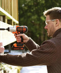 Black+Decker cordless drill with orange/black body drilling into wood; man wearing safety glasses.