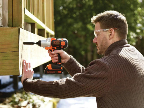 Black+Decker cordless drill with orange/black body drilling into wood; man wearing safety glasses.