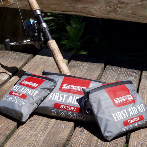 Pac-Kit Medical waterproof first aid kit pouches (Explorer 2/3) with red labels on wooden dock beside a fishing rod.