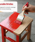 Hand using 3-inch paint brush on wooden stool, demonstrating smooth application, part of 6 Inch Deck Stain Brush With 77 Inch Extendable Handle And 3 Inch Paint Brush Set For Wood Fence And Wall Durable Synthetic Bristles For Smooth Even Deck Stain Application.