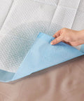 Hand holding a corner of Straight Up Reliable Disposable Underpads For Adults super absorbent incontinence bed pads on a surface.