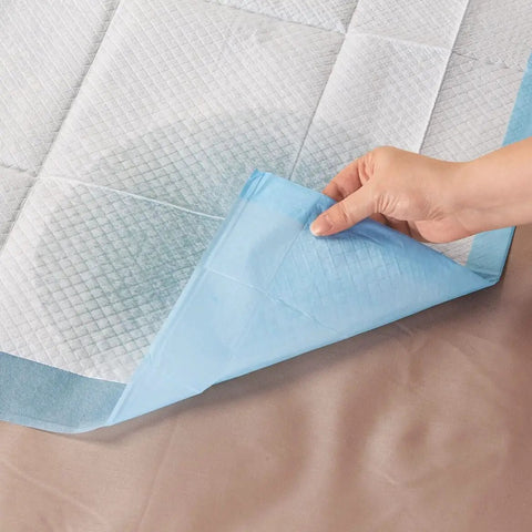 Hand holding a corner of Straight Up Reliable Disposable Underpads For Adults super absorbent incontinence bed pads on a surface.