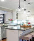 Modern kitchen with pendant lighting, featuring a tray with flowers and decor on a central island, stools, and white cabinetry.