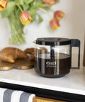 1.25L Glass Carafe for KBG Brewers, Clear Black Version, resting on kitchen counter next to croissants, holds 40 ounces of coffee