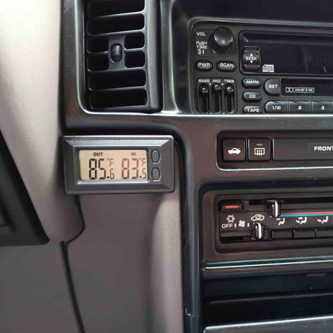 Black dual-display digital car thermometer mounted on the dashboard near the vent, showing outside and inside temperatures in Fahrenheit.