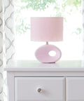 Pink ceramic table lamp with a round base and fabric shade on a white dresser in a bright room with patterned curtains.