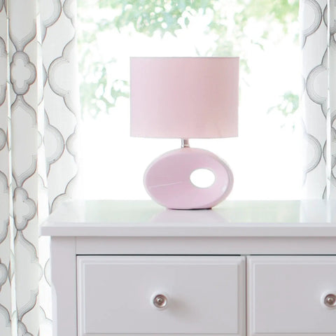 Pink ceramic table lamp with a round base and fabric shade on a white dresser in a bright room with patterned curtains.