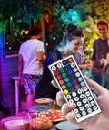 Hand holding 12V 44 Key Wireless IR Remote Controller With Dual Ports Rectifier Box And 3A Power Supply For 2835 3528 5050 RGB LED Strip Lights Flexible Tape at a party setting with LED lights.