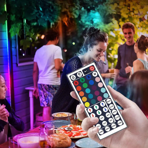 Hand holding 12V 44 Key Wireless IR Remote Controller With Dual Ports Rectifier Box And 3A Power Supply For 2835 3528 5050 RGB LED Strip Lights Flexible Tape at a party setting with LED lights.