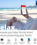 Person on beach holding phone showcasing YoLink smart sensor alert, illustrating home protection benefits.