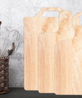 Four light wood charcuterie and cutting boards with handles standing upright against a wall next to a wicker basket with utensils.