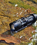 Ultra Bright Mini LED Flashlights 7W Zoomable Handheld Beams, Water Resistant, rugged, water-drenched flashlight on rocky surface.