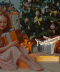 Lit 3D Airplane Illusion Night Light With Dimmable LED Wood Base beside a girl opening a gift near a Christmas tree
