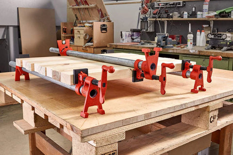 Bessey BPC-H34 3/4-in H-style pipe clamps in red, holding a wooden board on a workshop bench (angled view, five clamps).