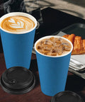 100 Pack 16 Oz Disposable To Go Coffee Cups With Lids, Sleeves And Stir Sticks For Grab And Go Hot Or Cold Drinks, Leakproof And Spill Resistant Paper Cups For Office Home Shop Events Navy with lids beside cups filled with hot coffee and iced coffee with a croissant on a tray in a cafe setting.