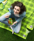 Top view of a woman sitting on a green picnic mat with salad, showing Tough As Nails 6 Inch Stainless Steel Patio Rug Stakes Set For Outdoor Use.