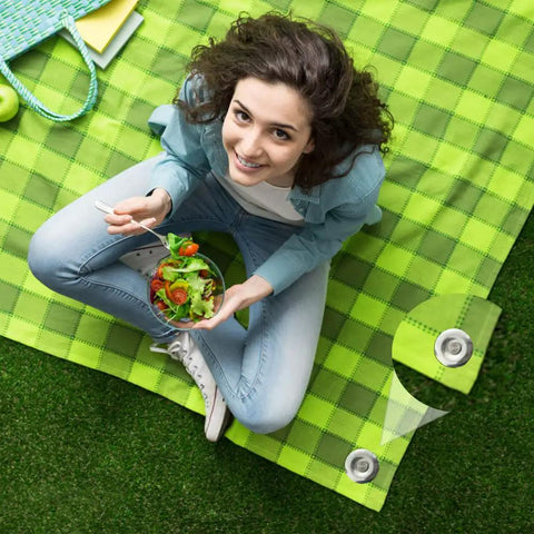Top view of a woman sitting on a green picnic mat with salad, showing Tough As Nails 6 Inch Stainless Steel Patio Rug Stakes Set For Outdoor Use.
