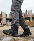 Person wearing Mens Steel Toe Work Boots Waterproof Safety Shoes For Job Sites, Rugged All Day Comfort, ASTM F2413-18 Certified, Black Industrial Boot With Durable Uppers stepping through mud on a construction site.