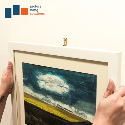 Person hanging framed artwork using heavy duty picture hangers for drywall and plaster 30 pound capacity 25 pack hardware.