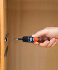 Hand holding Grab And Go 6V Cordless Screwdriver Kit With 14 Bit Set against wooden cabinet for quick home repairs.