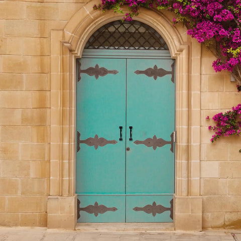 Turquoise double doors with iron strap hinges and handles set in a stone wall with purple flowers climbing on the side.