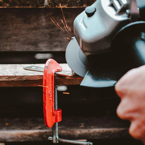 Angle grinder with a 4-1/2-inch flap disc grinding metal, sparks flying, secured in a red C-clamp vise.