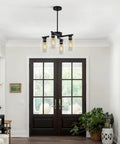Sleek Modern Black Four Light Farmhouse Chandelier Featuring Crisp Cylindrical Glass Shades Fully Adjustable Height Industrial Pendant Fixture above entrance.